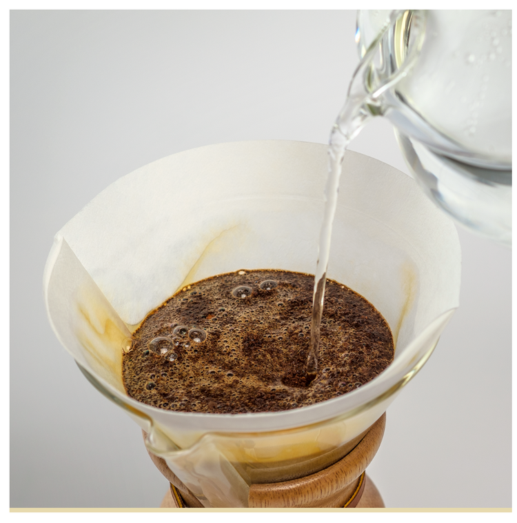 Et nærbillede af varmt vand, der hældes fra en glaskedel over malet kaffe i et papirfilter til en pour-over-brygning. Kaffemaskinen står på en træbase.