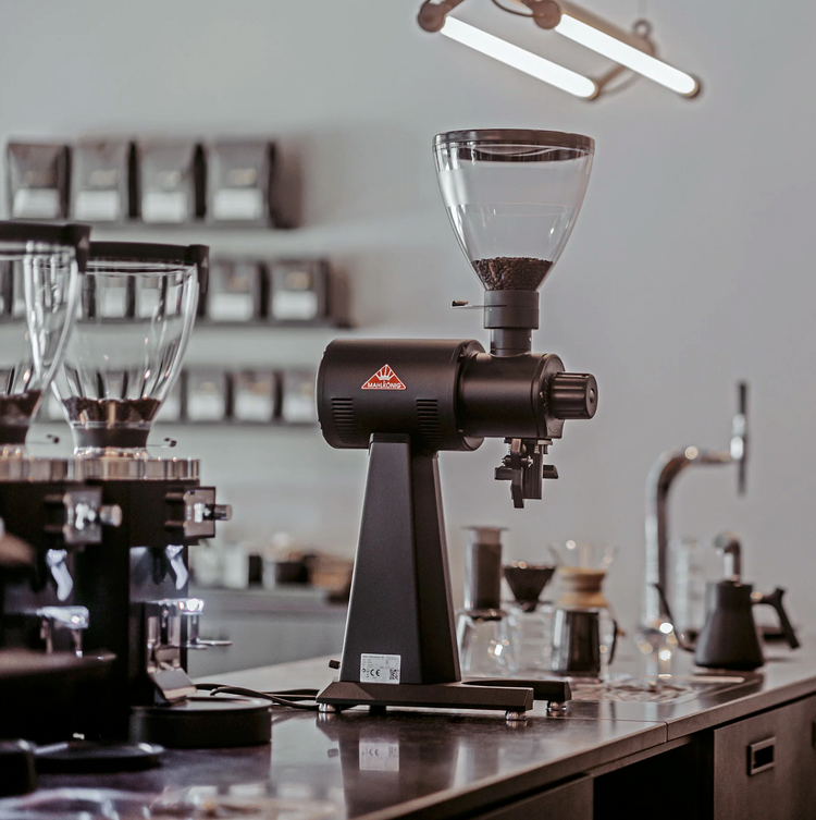 En elegant kaffekværn står på en disk i en moderne café, omgivet af kaffebryggerudstyr, krukker og hylder med kaffeposer i baggrunden.