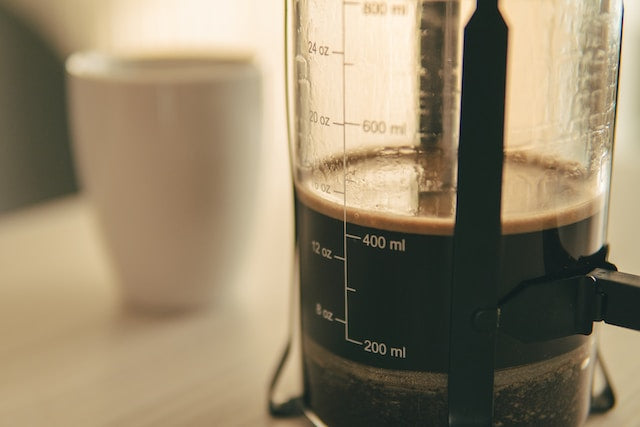 Nærbillede af en stempelkande med mørk kaffe, markeret med målestreger, og en sløret hvid kaffekop i baggrunden på en lys overflade.