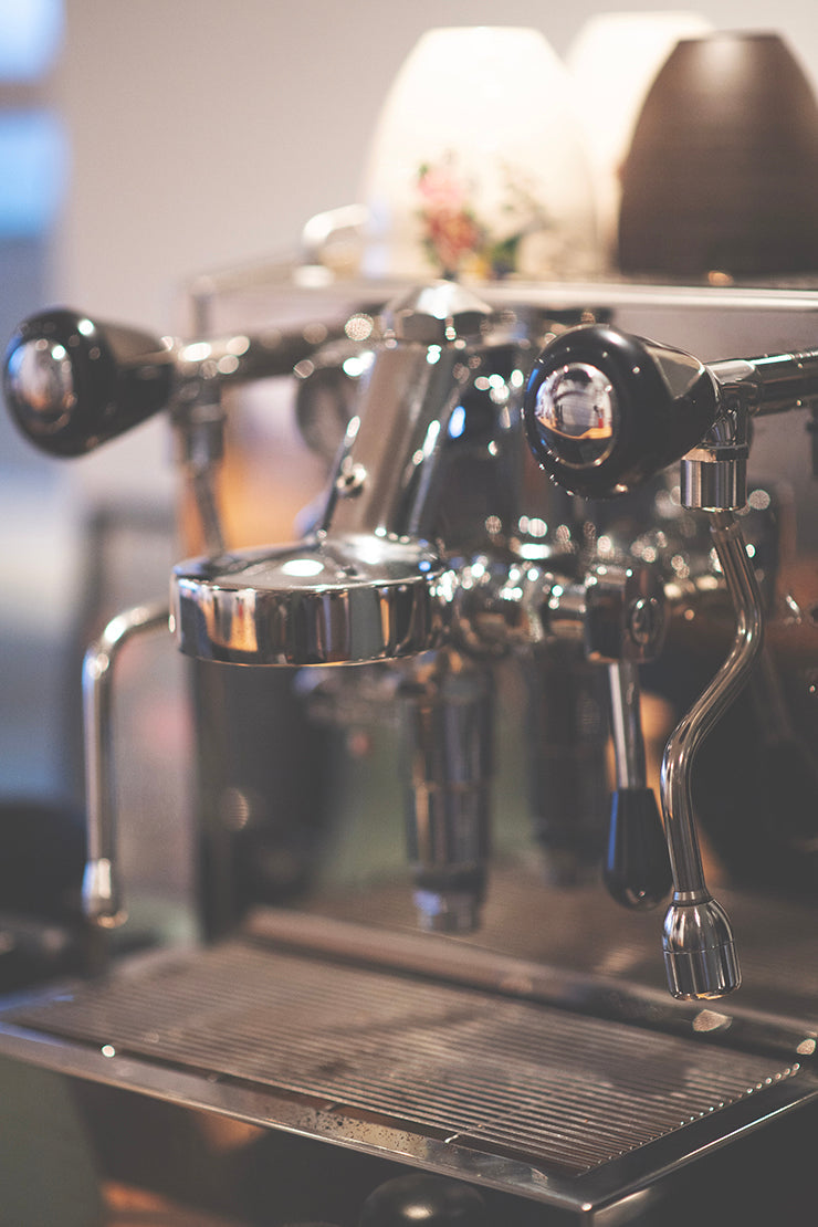 Et nærbillede af en skinnende espressomaskine med metalliske håndtag og en drypbakke. Slørede kopper er stablet oven på maskinen, og baggrunden er blødt ude af fokus.