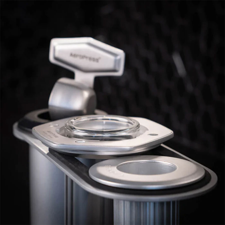 Nærbillede af Aeropress Premium Organizer fra Aeropress, lavet af rustfri stål med et bryggekammer i glas og et håndtag mærket "KEY", sat mod en mørk, tekstureret baggrund.