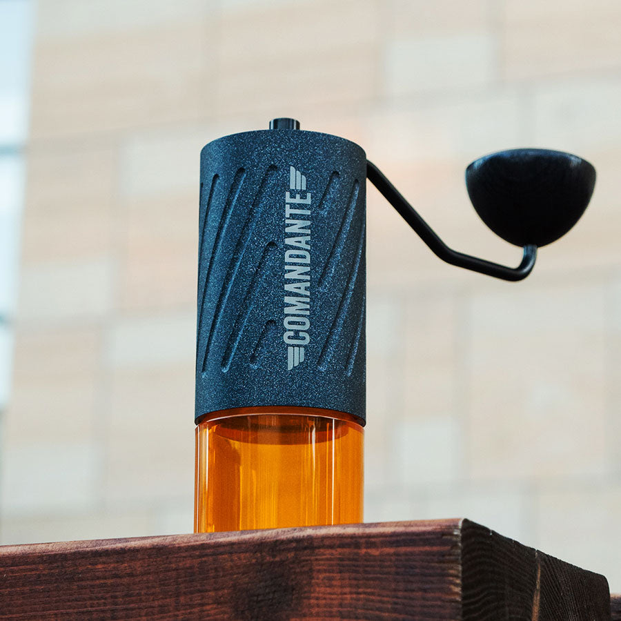 En Comandante C60 Baracuda - Nautilus Blue manuel kaffekværn hviler på en træoverflade og fremviser sin slående blå finish og robuste stålstruktur mod en blødt sløret beige flisebelagt baggrund.