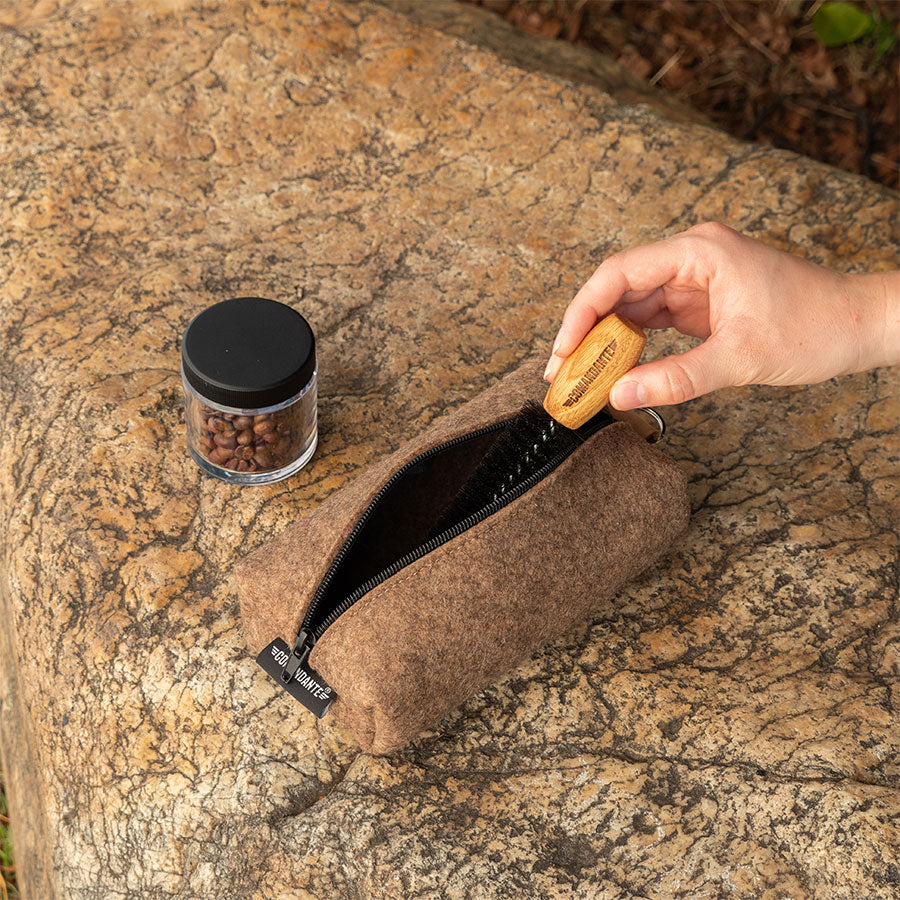 En hånd rækker ind i en brun, tekstureret pose på en stenet overflade, der ligner en Comandante Barista Værktøjstaske. Ved siden af er der en lille krukke med sort låg indeholdende kaffebønner, sat mod naturlige jordfarver.