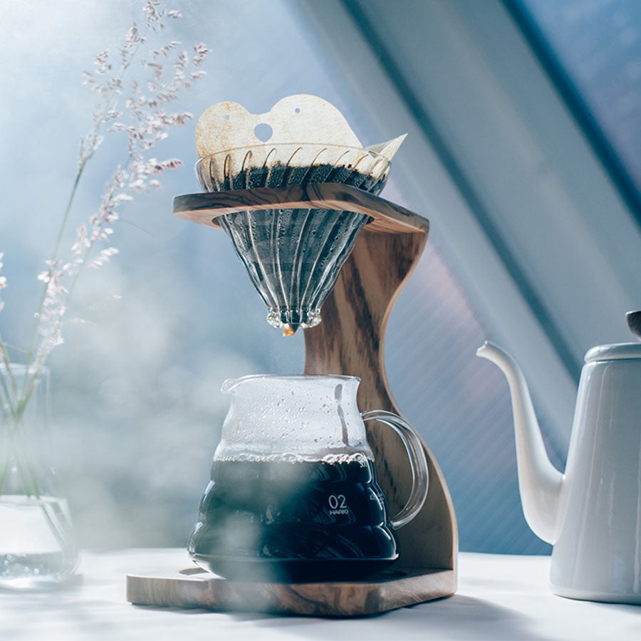 Sollyset fylder rummet, mens en Hario pour-over-kaffemaskine i glas med Hario Papir Filter Size 02 Love Birds brygger kaffe i en karaffel. I baggrunden er en tekande og en vase med fine stilke blødt oplyst.