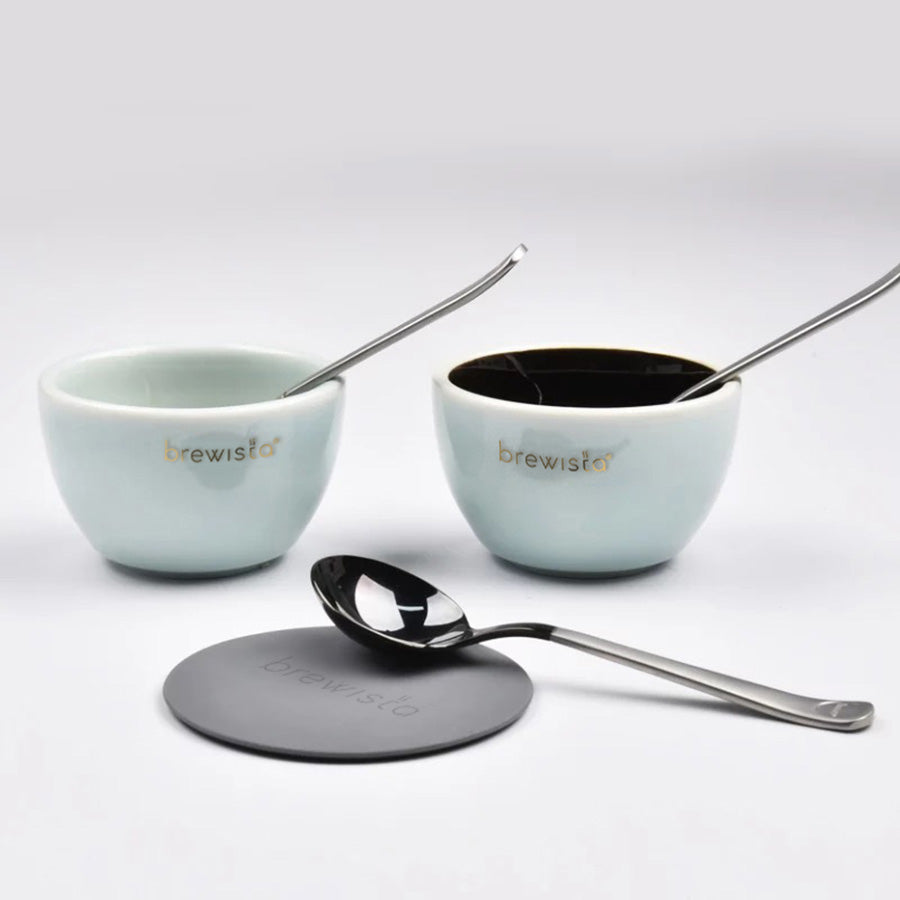To keramiske Brewista skåle mod en almindelig baggrund, hver ved siden af en slank Brewista Professional Cupping Spoon (Titanium - Sort). En slank sort underkop sidder foran. Skålene har et lyst ydre med mørkt interiør.