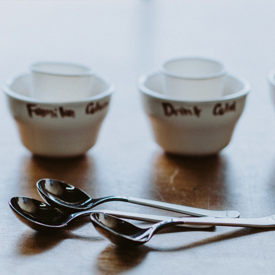 Fire hvide skåle med håndskrevne etiketter sidder på en træoverflade sammen med Brewistas Professional Cupping Spoon i titanium sort. I forgrunden er tre sølvskeer elegant arrangeret, som reflekterer lyset.
