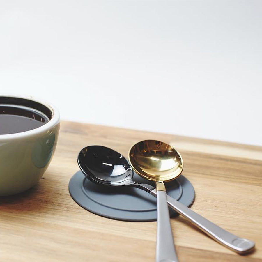 Et træbord viser en guld- og Brewista Professional Cupping Spoon (Titanium - Black), ideel til en Brewista-session. En kop mørk væske sidder på en rund, grå coaster, klar til ekspert-cupping ske-evaluering.