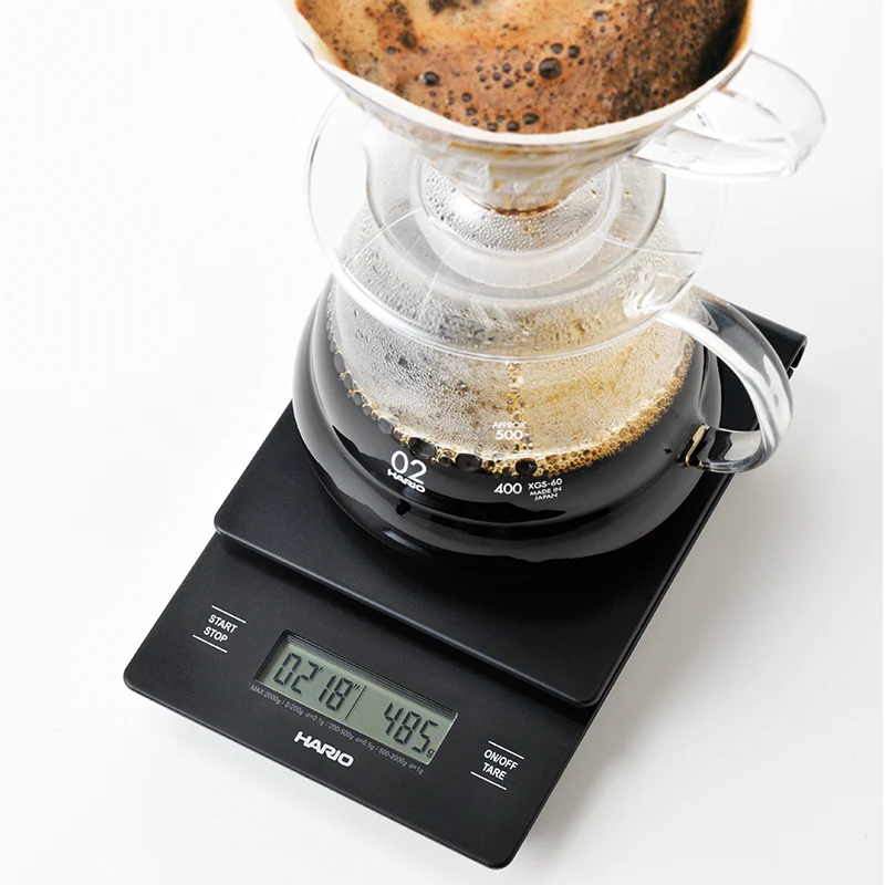 Hario V60 Drip Vægt har en klar drypper og karaffel i digital skala, der viser friskbrygget kaffe. Displayet viser måleenheder for tid og vægt, hvilket sikrer præcision i hver kop, der hældes over kaffe.