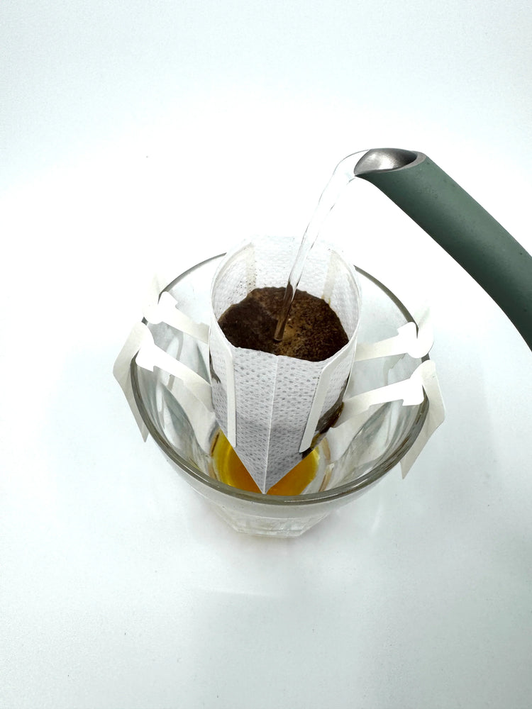 Varmt vand hældes i en dryp kaffepose over en glaskop, hvor brygget kaffe samles i bunden.