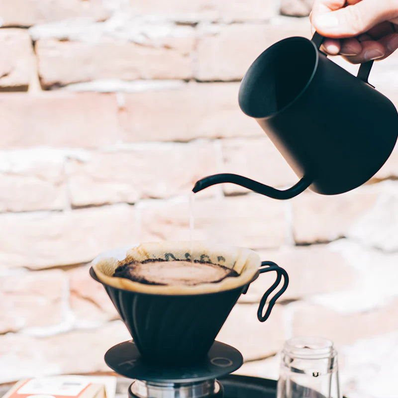 En hånd hælder varmt vand fra en sort kedel ned i en kaffedrypper med et papirfilter og laver pour-over-kaffe på en baggrund af en lys murstensvæg.