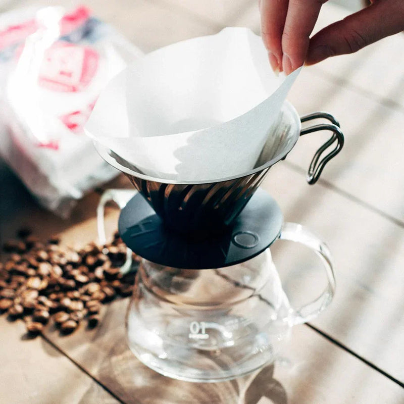 En hånd placerer et papirfilter i en kaffedrypper af metal, der står på en glaskaraffel, mens kaffebønner og en pose kaffe er synlige på en træplade i nærheden.