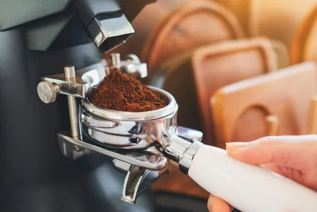 En hånd holder et portafilter fyldt med friskmalet kaffe under en kværn, klar til at lave espresso. Brunt kaffegrums er synligt, og der er slørede objekter i baggrunden.
