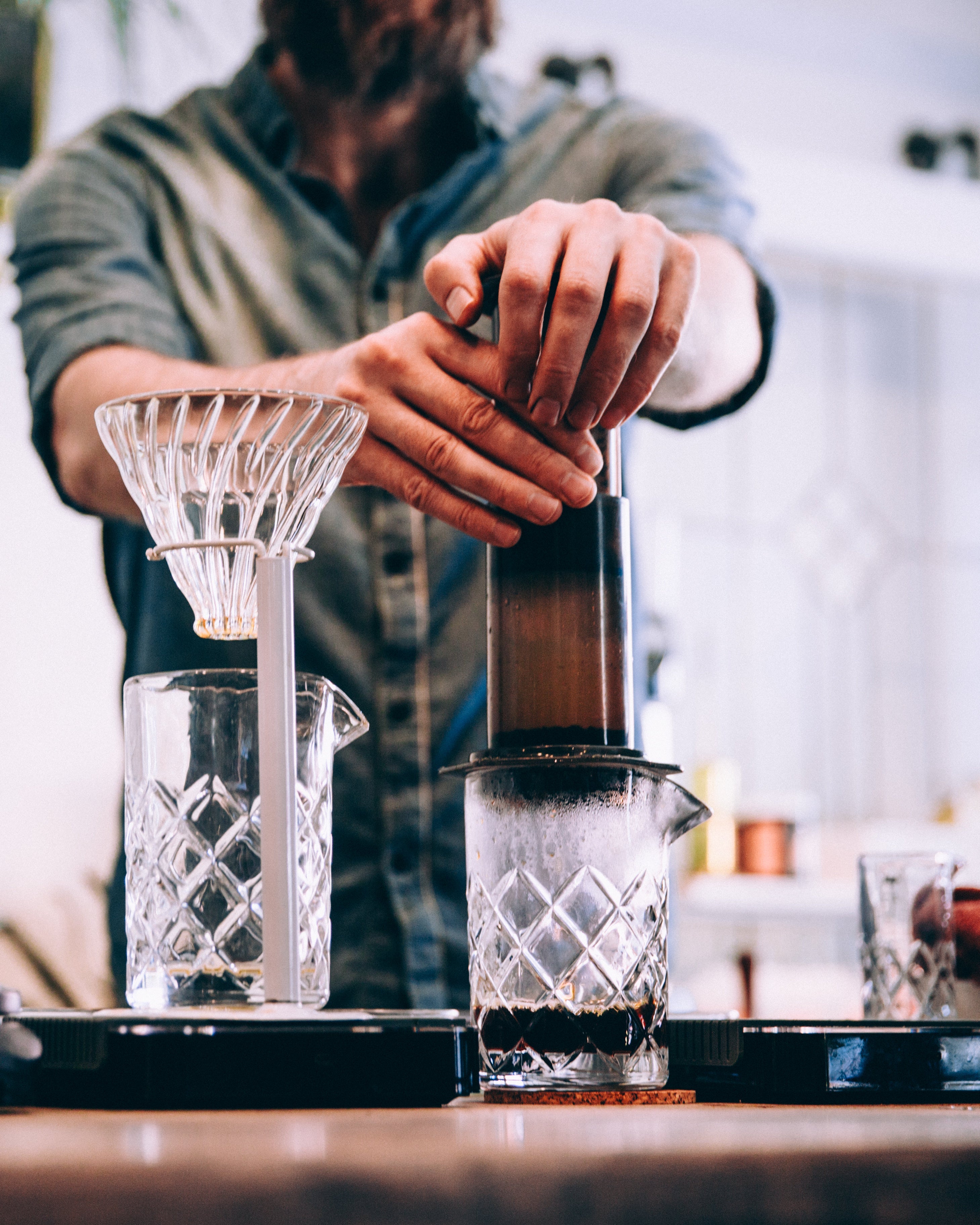 En person trykker på en AeroPress-kaffemaskine over en glasbeholder med et andet glas og en pour-over-kaffedrypper ved siden af på en træbordplade.