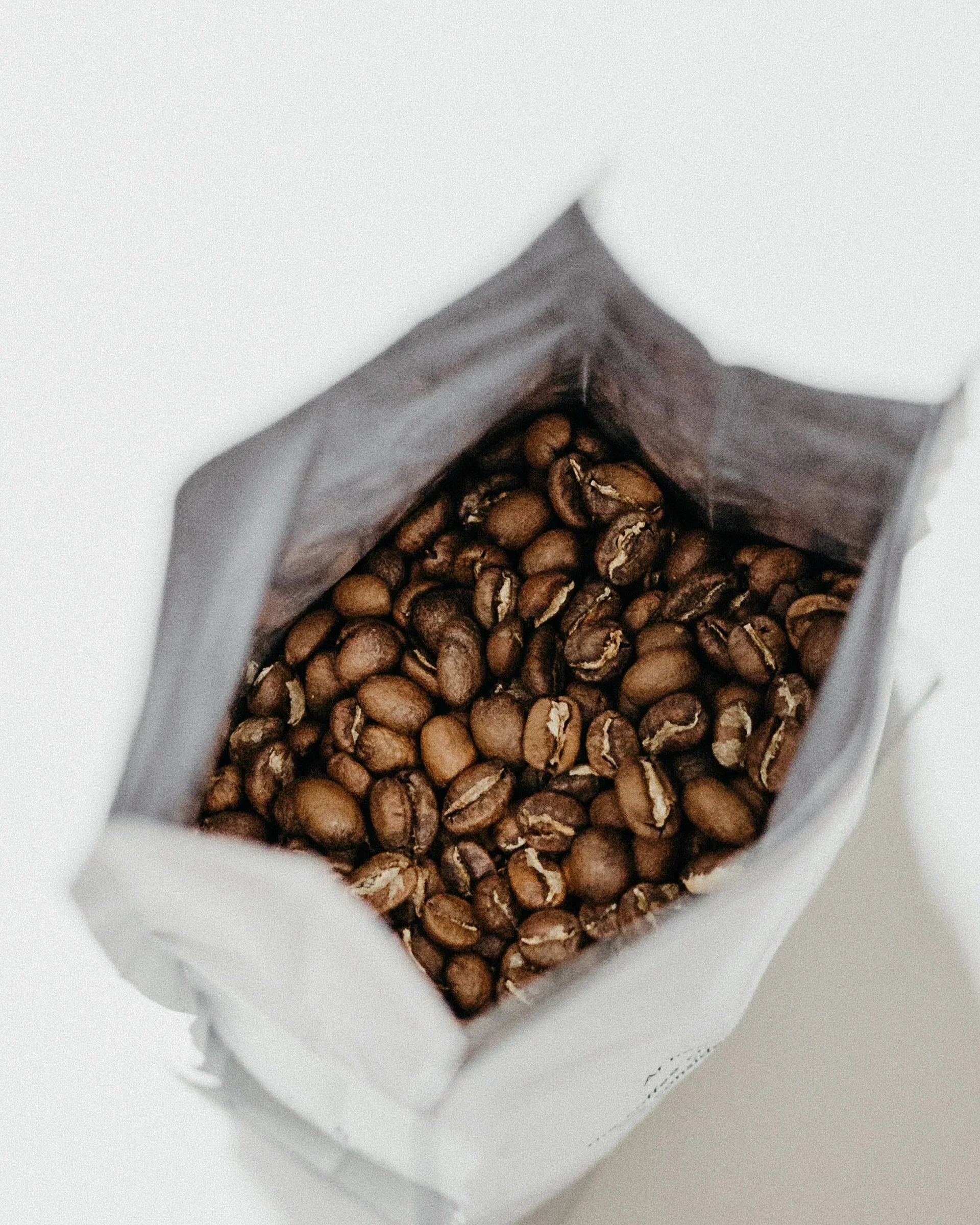 En åben hvid pose fyldt med ristede kaffebønner, set ovenfra mod en almindelig hvid baggrund.