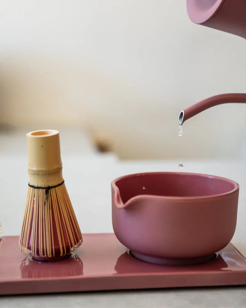 En lyserød tekande hælder vand i en matcha-skål i samme lyserøde farve ved siden af en bambus-matcha-pisker, og det hele står på et lyserødt bakke foran en neutral baggrund.