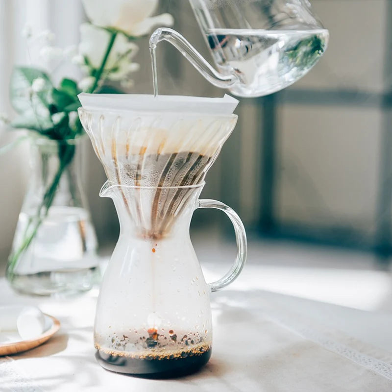 En glaskedel hælder varmt vand over kaffen i en Hario Kaffe Decanter 400 ml pour-over-maskine, med blomster og sollys i baggrunden på et lyst bord.