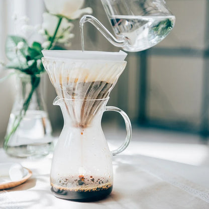 En glaskedel hælder varmt vand over kaffen i en Hario Kaffe Decanter 400 ml pour-over-maskine, med blomster og sollys i baggrunden på et lyst bord.