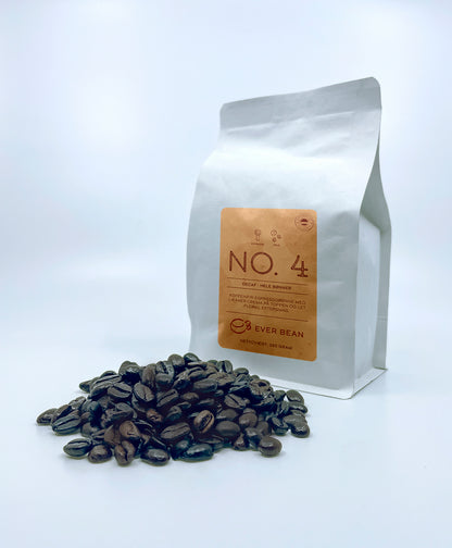 En hvid EverBean-pose mærket "No. 04 · Colombia Decaf, Whole Beans" dukker op ved siden af en bunke mørkristede kaffebønner med branding og vægtdetaljer på en almindelig hvid baggrund.