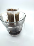En EverBean ApS. CoTa Blend · Single Serve Drip Bag med vådt kaffepulver oven på et glas, hvor der brygges frisk kaffe, som samler sig i bunden af glasset på en hvid baggrund.