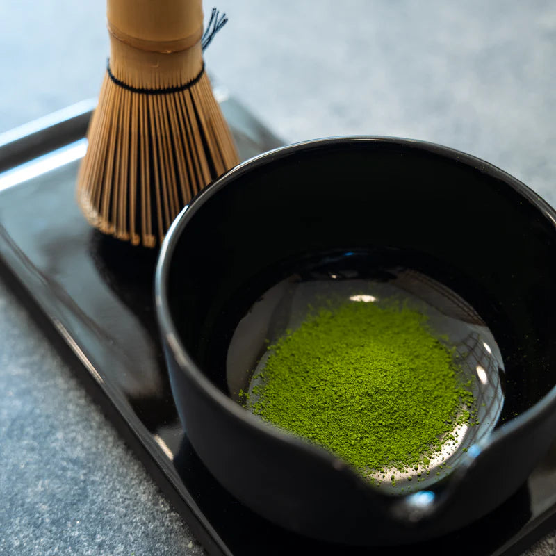 En sort skål med strålende grøn matcha og en bambuspisk fra Fellow Matcha-sættet – Komplet Matcha-sæt fra Fellow – står på et sort bakke oven på en grå bordplade.