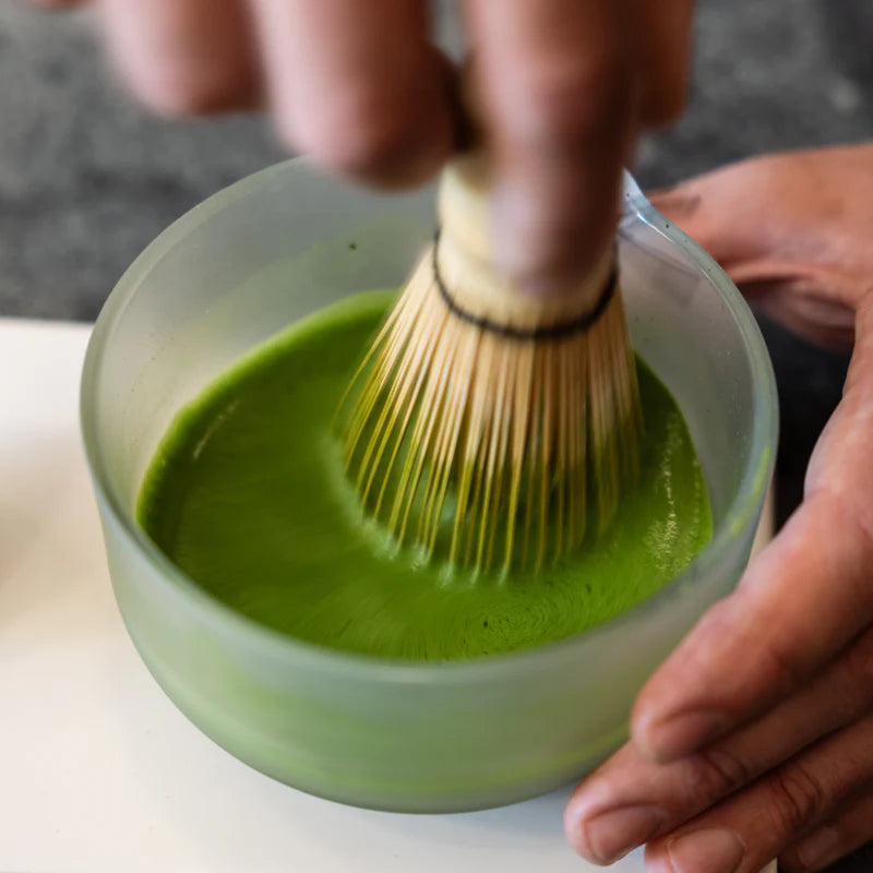 En hånd bruger et bambuspiskeris fra Fellow Matcha Set – Komplet Matcha Sæt fra Fellow til at piske den strålende grønne matcha-te i en gennemsigtig skål, så der dannes et skummende lag på toppen.