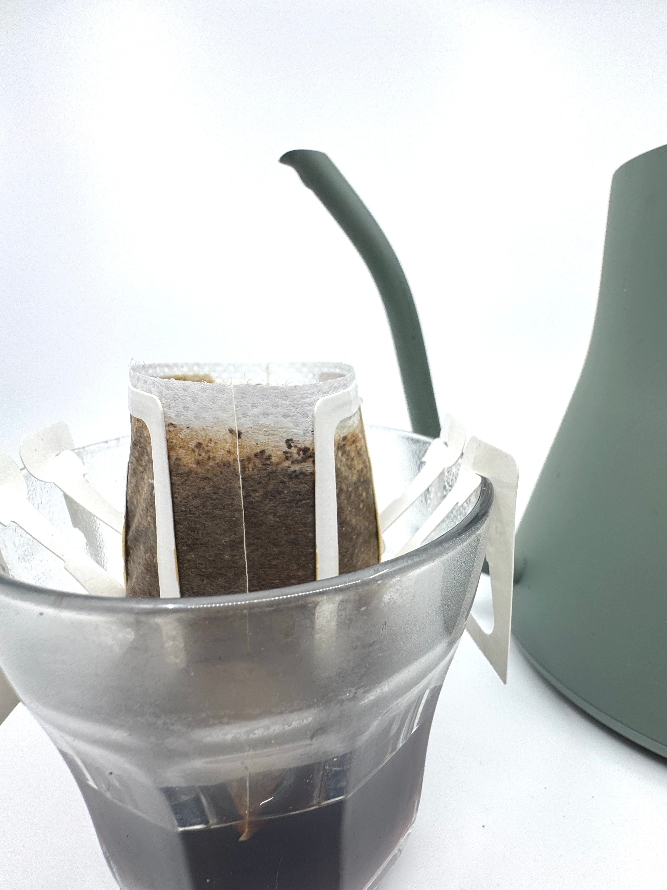 Et Single Serve, No. 08 - Etiopien kaffefilter fra EverBean ApS. sidder på toppen af en glaskop, mens varmt vand strømmer ud af en grøn svanehals-kedel på en almindelig hvid baggrund.