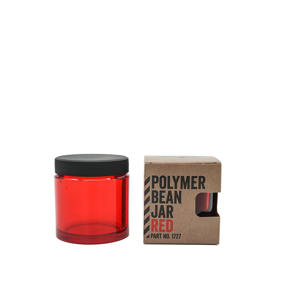 En Comandante Polymer Bean Jar med låg i rød, kompatibel med C40 og toppet med et sort låg, sidder ved siden af en brun æske. Æsken viser "Polymer Bean Jar Red" og "Part No. 1727," med en udskæring, der afslører en del af krukken indeni.