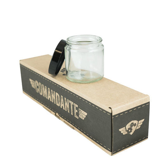 Et lille, klart kaffebønne glas fra Comandante Klar 4 pak, med lufttætte design og sort låg, sidder på en brun-sort Comandante æske. Kassen, ideel til enhver kaffeentusiast, har et logo med vinger og et geardesign.