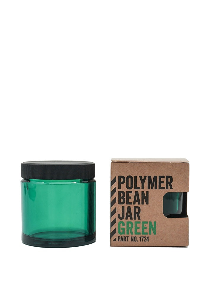 En grøn gennemsigtig polymerkrukke med sort låg, mærket "Comandante, Polymer Bean Jar med låg, Grøn," er udstillet ved siden af en brun papkasse, der har mærkenavnet Comandante og har en udskæring, der afslører en del af glasset indeni.