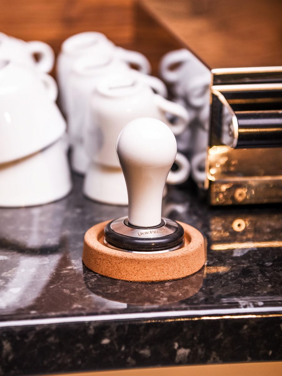 En Joe Frex Stamper Station, Kork, med en træbase hviler på en marmorbordplade. Bag den er flere stablede hvide kaffekopper og en metallisk espressomaskinedel, delvist synlig.