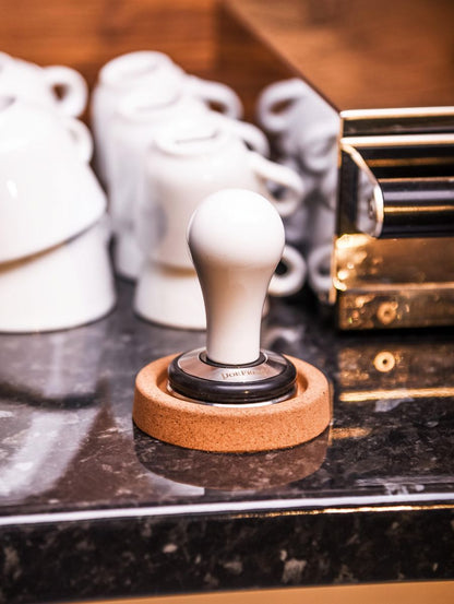 En Joe Frex Stamper Station, Kork, med en træbase hviler på en marmorbordplade. Bag den er flere stablede hvide kaffekopper og en metallisk espressomaskinedel, delvist synlig.