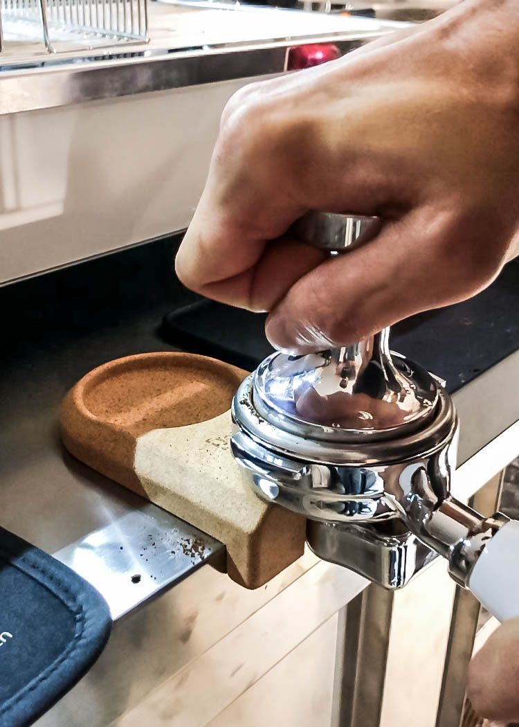 En hånd tamper dygtigt kaffegrums i et portafilter ved hjælp af en metaltamper på en disk. Under hviler Joe Frex Stamper Station, Kork, en miljøvenlig måtte lavet af genanvendt kork for stabilitet og bæredygtighed.