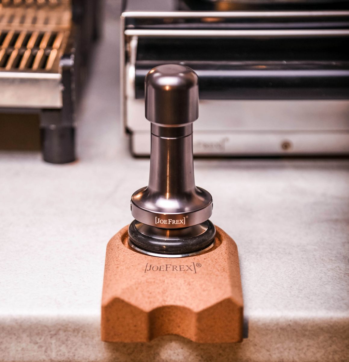 En Joe Frex metallisk kaffetamper sidder på Stamper Station Kork-holderen på en bordplade. Bagved fremhæver slørede espressomaskinedele et økologisk produkt, der kombinerer funktionalitet med miljøvenligt design.