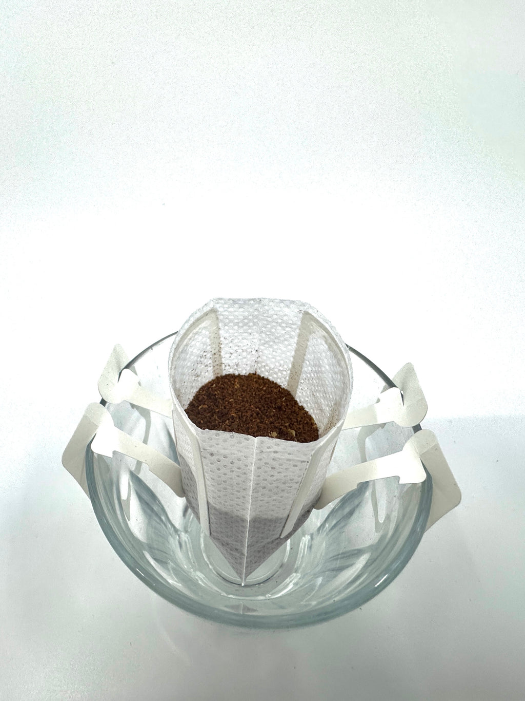 En glasskål holder et drypkaffefilter af EverBean ApS. Single Serve, No. 04 - Colombia, Decaf (øko). Filterets arme hænger over skålens kant, så det er nemt at brygge, vist mod en hvid baggrund.