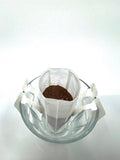 En nr. 4 · Colombia Decaf · Øko · Single Serve Drip Bag fra EverBean ApS. ligger over et gennemsigtigt glas, klar til at blive hældt varmt vand over, på en ensfarvet hvid baggrund.