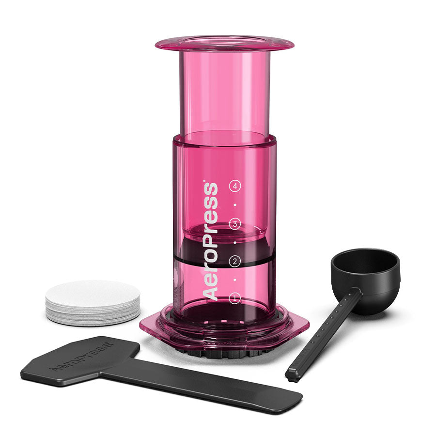 AeroPress, Limited, Kaffebrygger by Aeropress i pink indeholder en scoop, omrører, papirfiltre og en filterholder. Den har innovativ bryggeteknologi og er vist på en hvid baggrund.