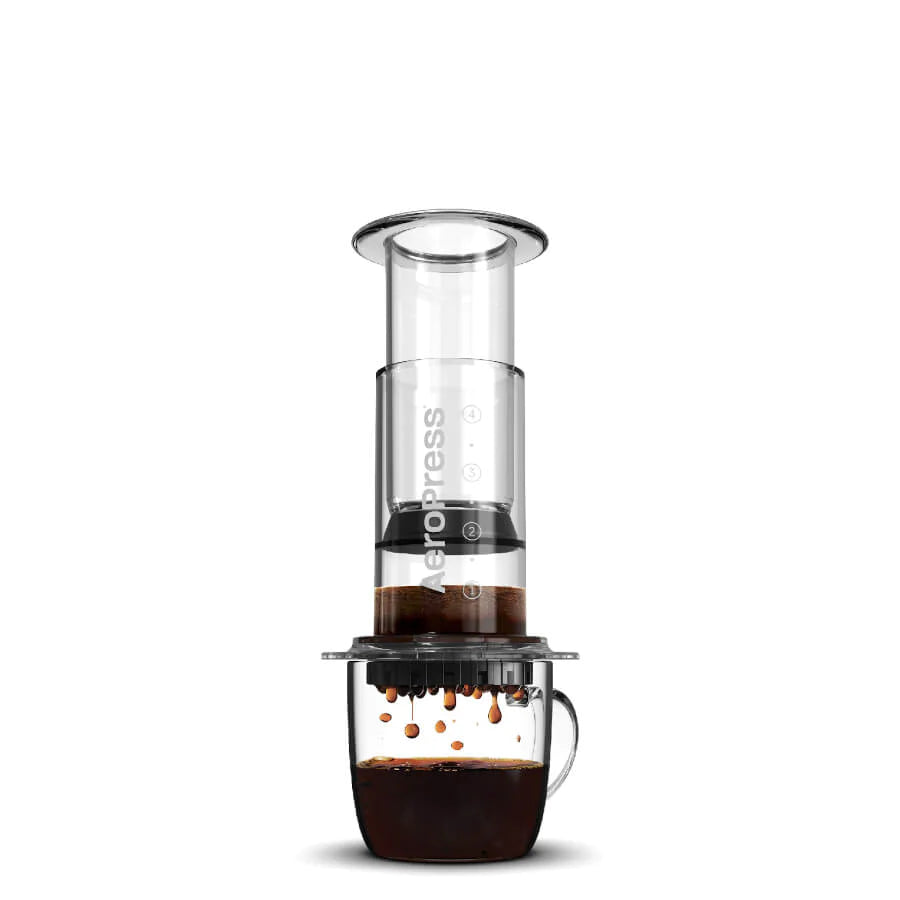 En AeroPress, Clear, Kaffebrygger by Aeropress hviler på et glaskrus, mens kaffen filtreres ned i kruset. Baggrunden er hvid og ren.