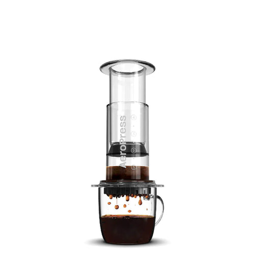En AeroPress, Clear, Kaffebrygger by Aeropress hviler på et glaskrus, mens kaffen filtreres ned i kruset. Baggrunden er hvid og ren.