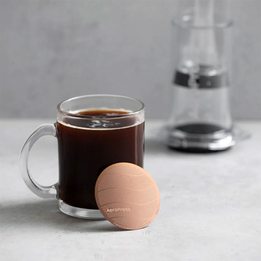 Et klart glaskrus med sort kaffe står på en grå overflade med AeroPress Gold Tone Filter fra Aeropress foran og en sløret kaffemaskine i baggrunden.