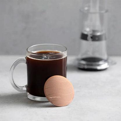 Et klart glaskrus med sort kaffe står på en grå overflade med AeroPress Gold Tone Filter fra Aeropress foran og en sløret kaffemaskine i baggrunden.
