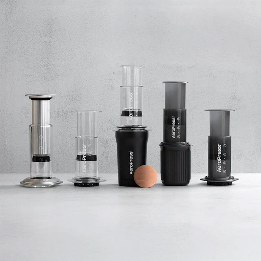 Seks AeroPress-kaffemaskiner og tilbehør, herunder Aeropress Gold Tone Filter, er stillet op på en lysegrå overflade mod en minimalistisk, lysegrå baggrund.