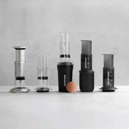 Seks AeroPress-kaffemaskiner og tilbehør, herunder Aeropress Gold Tone Filter, er stillet op på en lysegrå overflade mod en minimalistisk, lysegrå baggrund.