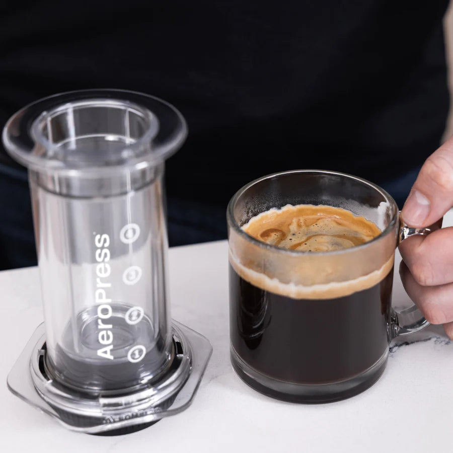 En person i en sort skjorte holder et glas kaffe ved siden af en tom, opretstående AeroPress Clear Kaffebrygger fra Aeropress, lavet af slidstærkt Tritan™-materiale på bordpladen.