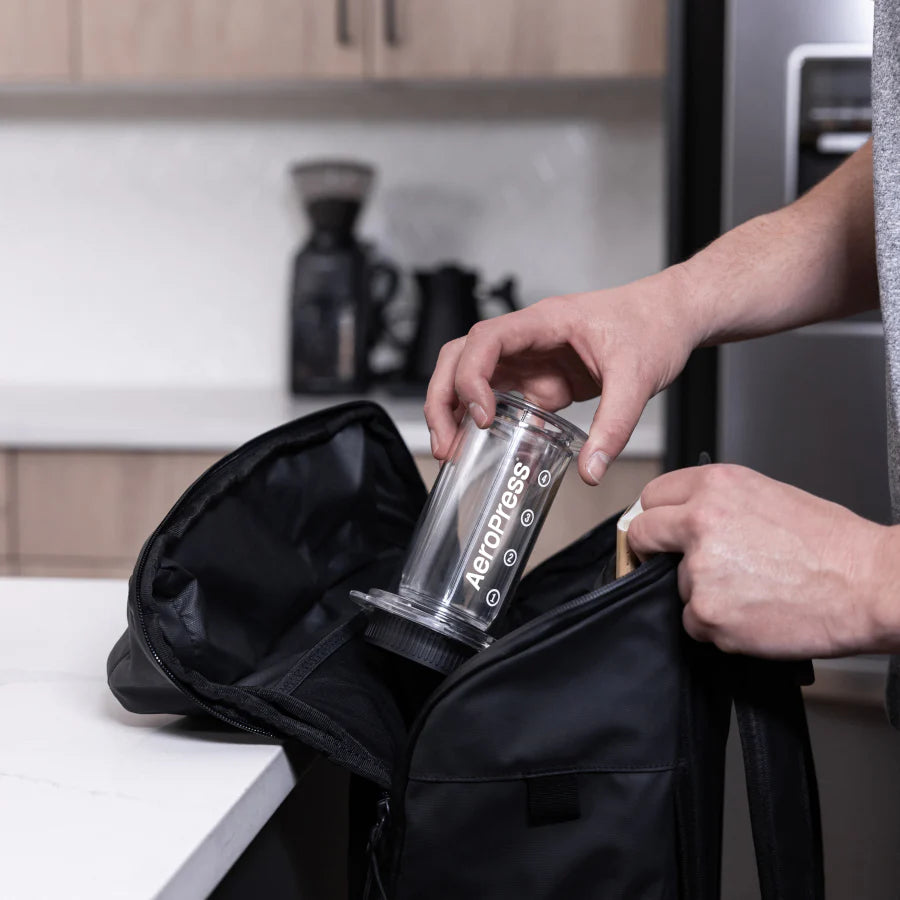 Nogen placerer en AeroPress Clear Kaffebrygger fra Aeropress i en sort rygsæk på en køkkenbord. Scenen byder på træskabe og et subtilt apparat i baggrunden.