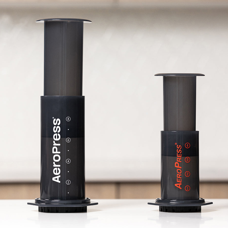 Aeropress XL kaffebrygger og den kompakte AeroPress Go står side om side på en hvid flade, med en uskarp lys baggrund.