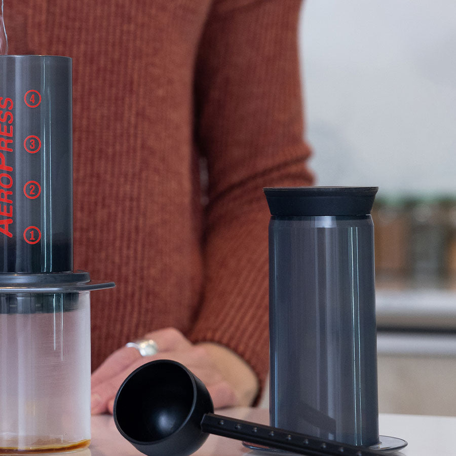 En person i en brun sweater fremviser deres kaffebrygningsevner ved siden af en delvist samlet Aeropress på køkkenbordet, med det pænt arrangerede adskilte gummistempel, kammer og scoop.