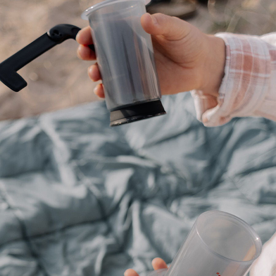 En person i en plaid skjorte holder en Aeropress gummistempel kaffekværn over et tæppe udendørs, med den klare plastikkværns sorte håndtag og base hjulpet af en anden hånd nedenunder.