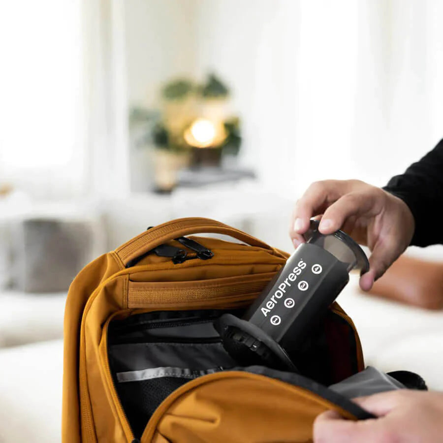 En person pakker forsigtigt en EverBean Aeropress Barista Pack og en pose kaffebønner i en orange rygsæk. I den slørede baggrund skaber en hyggelig sofa og en frodig plante den perfekte ramme om et koffeindrevet eventyr.