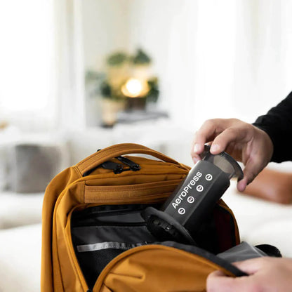 En person pakker forsigtigt en EverBean Aeropress Barista Pack og en pose kaffebønner i en orange rygsæk. I den slørede baggrund skaber en hyggelig sofa og en frodig plante den perfekte ramme om et koffeindrevet eventyr.