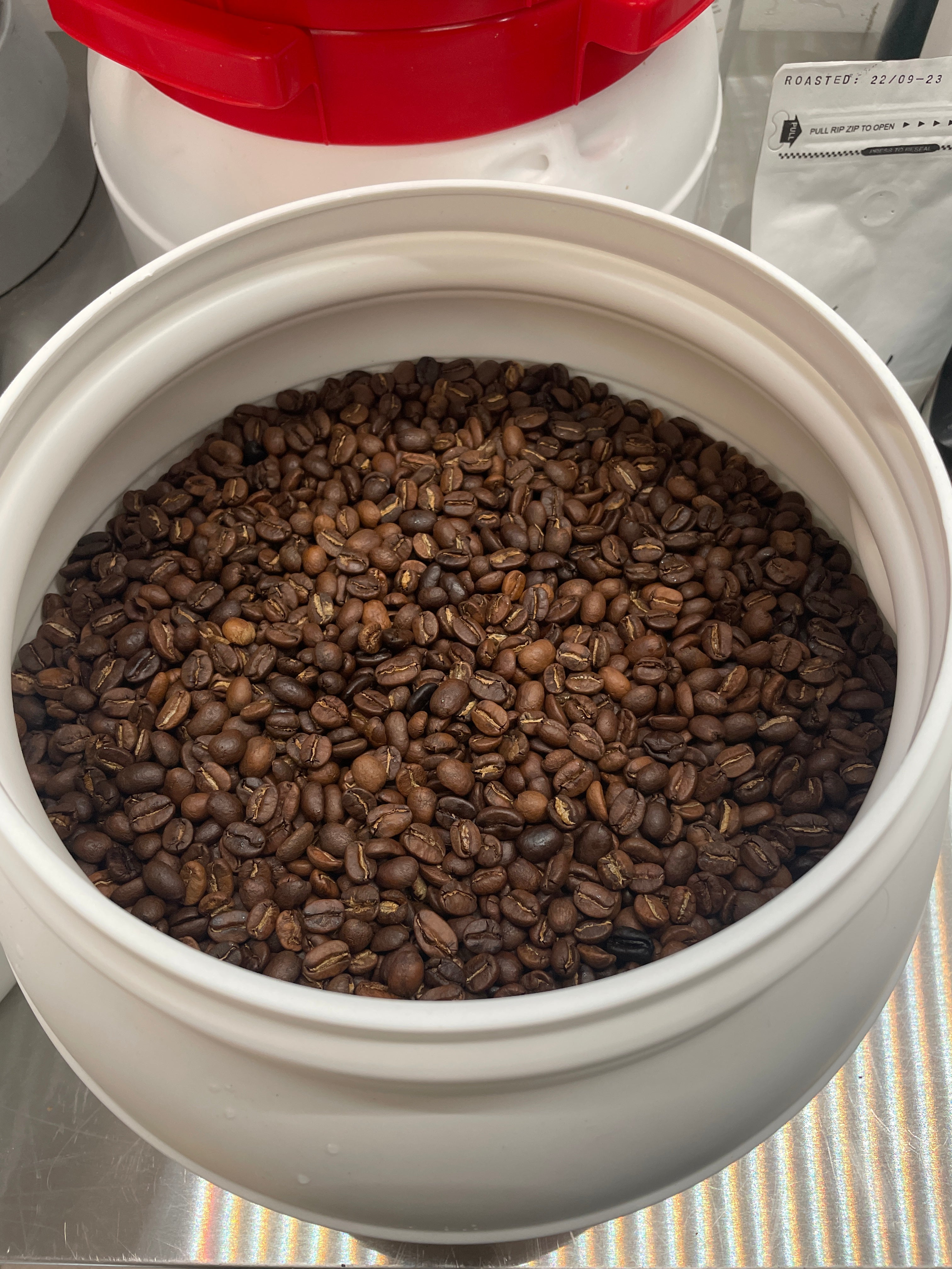En stor hvid EverBean Limited ICED Coffee Blend (w) beholder fyldt med mørkristede Arabica-bønner står på rustfrit stål. I baggrunden er en beholder med rødt låg og en pose koldbrygget kaffe synlige.